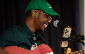 Young man in green ball cap, playing guitar and singing into a microphone.